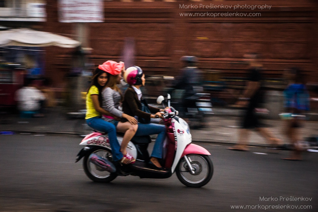 Three girls speeding by on a pinky-white scooter